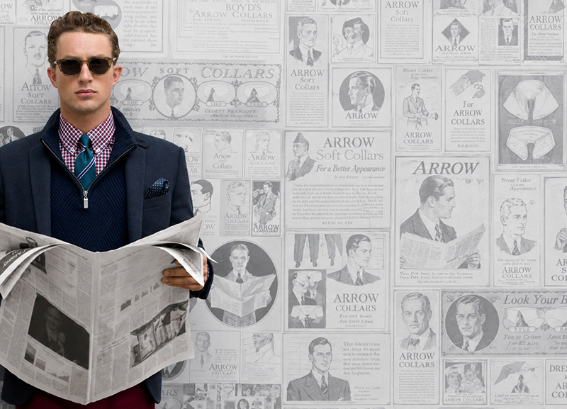 Mannequin ARROW tenant un journal devant une fresque d’archives de la marque